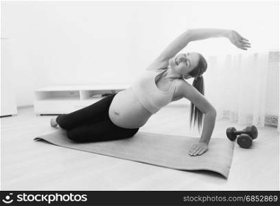 Black and white image of beautiful pregnant woman stretching on fitness mat at home