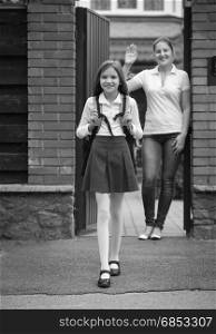 Black and white image of beautiful smiling girl in schoolgirl standing in front of mother waving to her