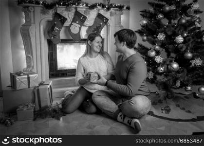 Black and white image of handsome young man sitting at fireplace with woman and giving her Christmas present