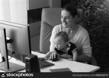 Black and white image of happy smiling woman working at computer with her little baby son