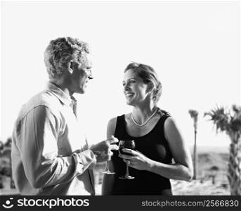 Black and white image of mid-adult Caucasian couple holding wine glasses and smiling at each other.