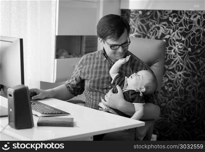 Black and white image of smiling young man working in home office and looking after his baby son. Black and white photo of smiling young man working in home office and looking after his baby son