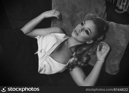 black and white image of very cute and sweet blond young woman , laying on the sofa , she has elegant dress , and long hair . she is looking up with a dreaming expression