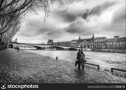 Black and white image with the Seine River, its shore where people walk, an old bridge and the Orsay Museum, under a dramatic sky, on a chilly day of February.