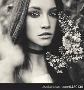 Black and white outdoor fashion photo of beautiful young woman surrounded by flowers of lilac. Spring blossom