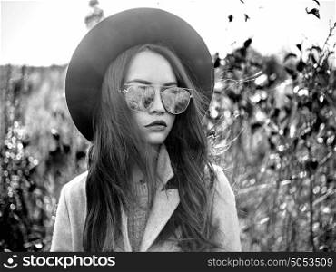 Black and white Outdoor fashion photo of young beautiful lady in autumn landscape with dry flowers. Gray coat, black hat, sunglusses, wine lipstick. Warm Autumn. Warm Spring