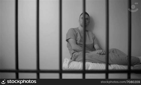 Black and white pensive inmate sits on bed in prison