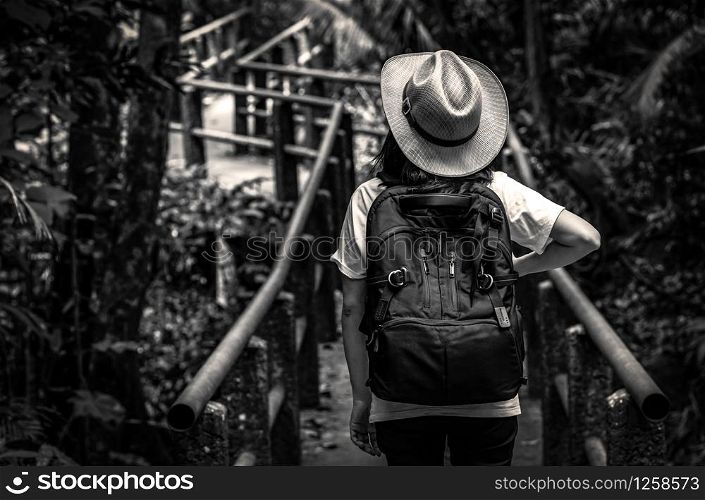 Black and white photo of Asian woman tourist with hat and backpack standing and start walking on nature trail bridge in evergreen forest. Alone young woman traveler.