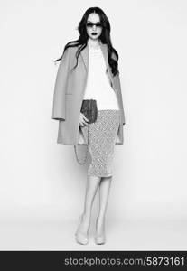 Black and white photo of beautiful brunette model in fashion clothes posing in studio. Wearing coat, handbag, shoes