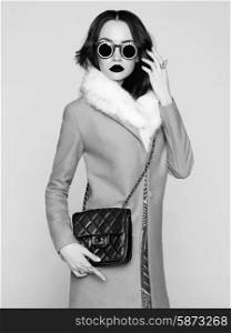 Black and white photo of beautiful brunette model in fashion clothes posing in studio. Wearing coat, handbag, sunglasses