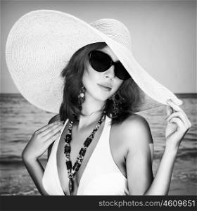 Black and white photo of beautiful female posing on the beach, wearing stylish big hat and sunglasses, luxury summer vacation concept