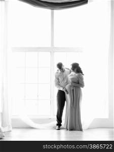 Black and white photo of elegant man hugging pregnant wife against big window