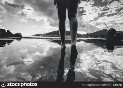 Black and white photo of female legs walking on water with sky reflecting on it