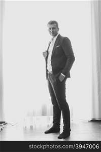 Black and white photo of handsome stylish man in suit posing against big window at modern interior