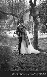 Black and white photo of happy bride and groom hugging under tree at autumn park
