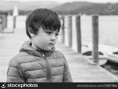 Black and white photo of sad kid standing alone by the lak with blurry pier background, Lost child looking down and crying,Lonely boy with unhappy face. Spoiled children concept