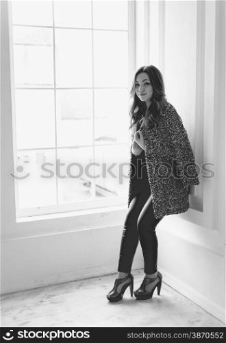 Black and white photo of stylish woman in leggings and jacket leaning against white wall