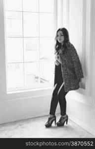 Black and white photo of stylish woman in leggings and jacket leaning against white wall