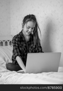 Black and white photo of teenage girl sitting on bed and using laptop