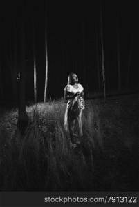 Black and white photo of woman in white dress walking at forest at night with lamp