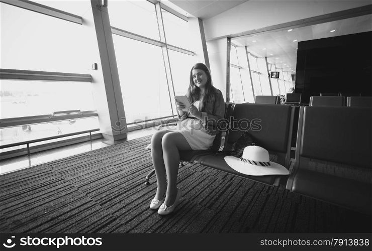 Black and white photo of young woman sitting in airport terminal and using tablet