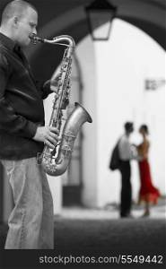 Black and white photograph street musician playing his saxophone while a romantic couple can be seen out of focus in the background the woman is in color wearing a red dress