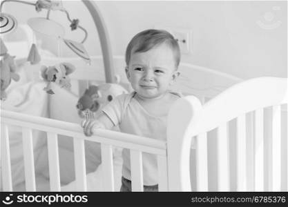 Black and white portrait of baby boy standing in crib and crying