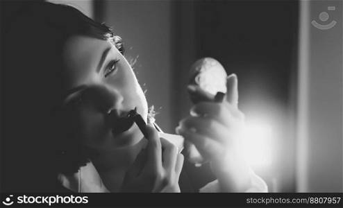 Black and white portrait of beautiful young woman applying lipstick while looking in small mirror. Retro or vintage style. Attractive brunette girl.