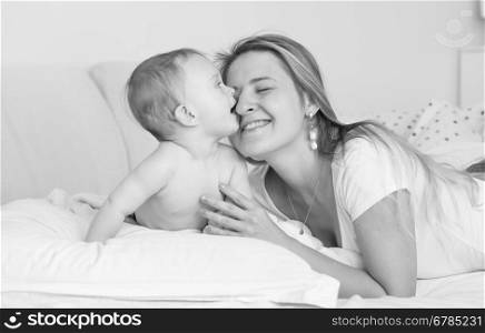 Black and white portrait of cheerful mother hugging and kissing her baby boy on bed