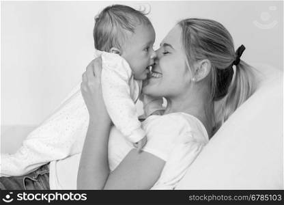 Black and white portrait of cheerful young mother lying on bed and kissing her 3 months old baby