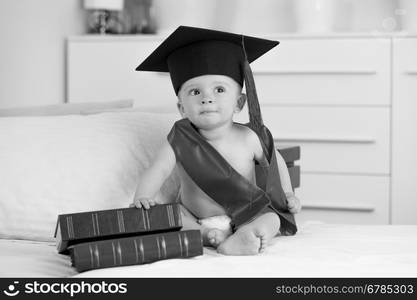 Black and white portrait of cute baby boy in graduation sitting with books on sofa