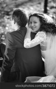 Black and white portrait of cute smiling bride hugging grooms back while sitting on leaves at park
