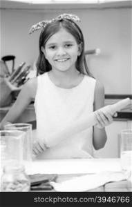 Black and white portrait of cute smiling girl holding wooden rolling pin while making dough