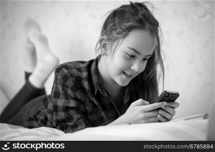 Black and white portrait of cute teenage girl lying on bed and typing message on phone