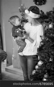 Black and white portrait of happy young mother in Santa hat and her 10 months old baby son posing at Christmas tree in living room