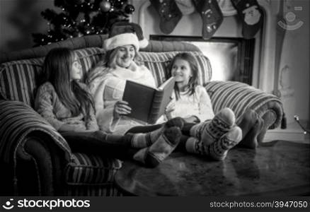 Black and white portrait of mother with two daughter reading book on sofa at fireplace