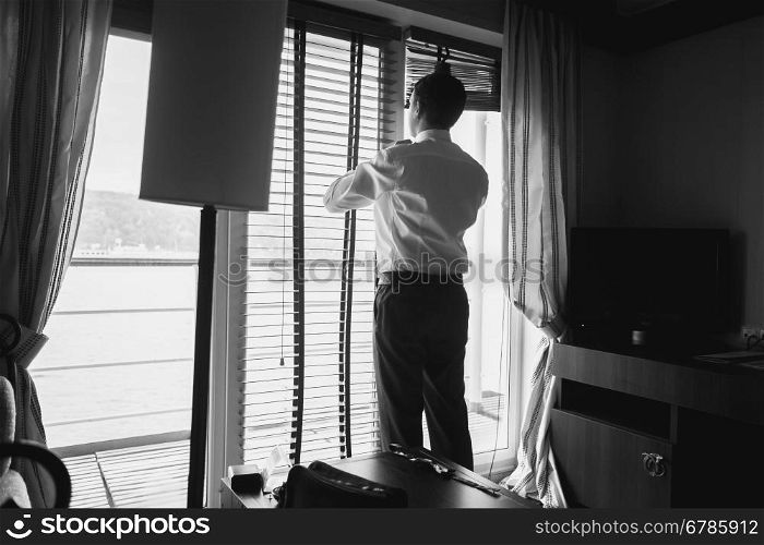 Black and white portrait of stylish businessman looking out of window at hotel room