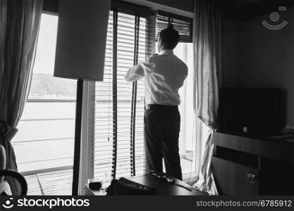 Black and white portrait of stylish businessman looking out of window at hotel room