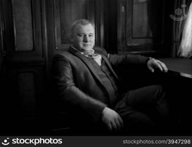 Black and white portrait of stylish man in retro suit sitting in armchair at office