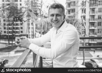 Black and white portrait of stylish smiling man in white shirt posing on balcony against urban background