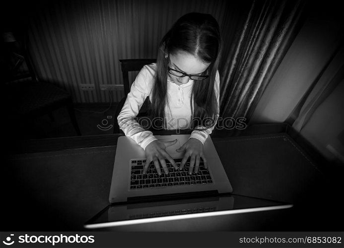 Black and white portrait of teenage girl sitting in dark room with laptop