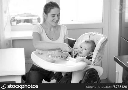 Black and white portrait of young smiling mother feeding her baby from spoon on kitchen