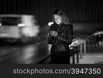 Black and white portrait of young woman typing message on street at night