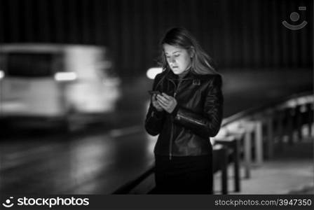 Black and white portrait of young woman typing message on street at night