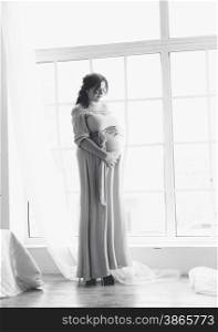 Black and white shot of beautiful pregnant woman posing against big window