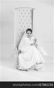 Black and white shot of elegant bride posing on white armchair at studio
