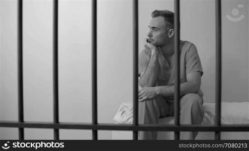 Black and white view of dejeacted inmate sitting behind bars in prison