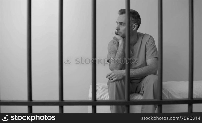 Black and white view of downcast inmate sitting behind bars in prison