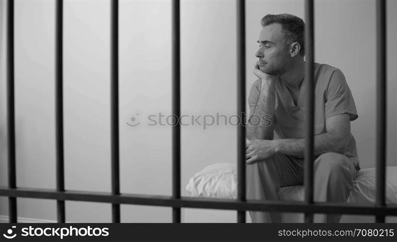 Black and white view of forlorn inmate behind bars in prison