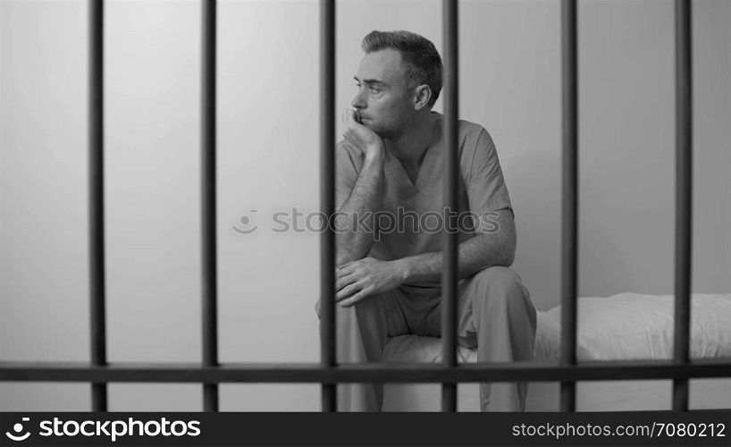 Black and white view of mournful inmate sittting on bed in prison